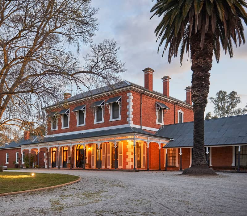 A grand historic brick mansion at dusk with lit porches and a tall palm tree.