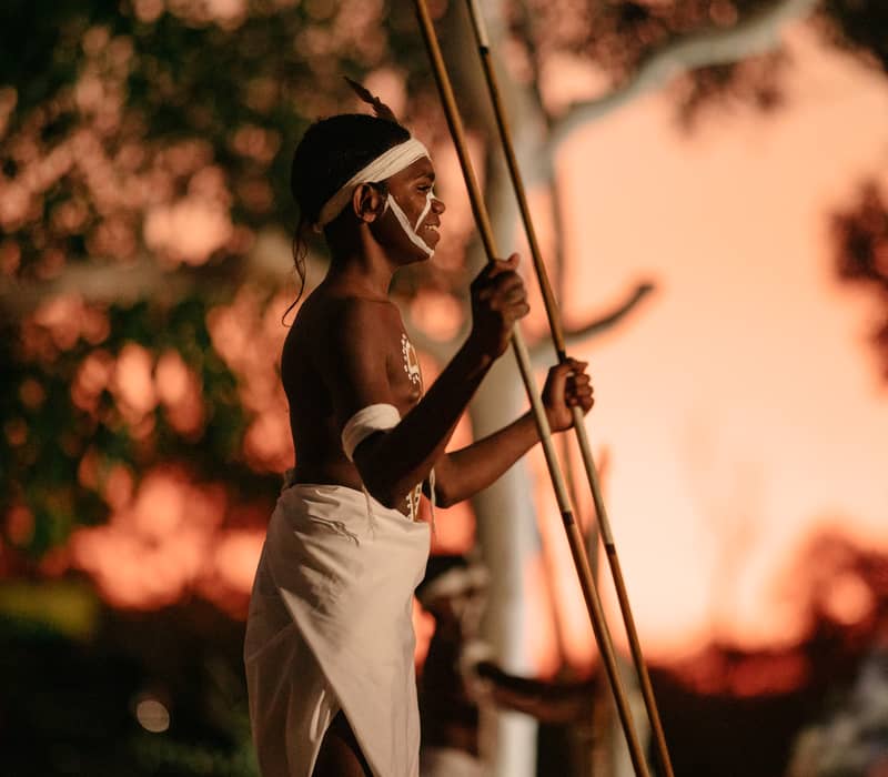 A profile shot of an Indigenous man with body paint holding spears, captured at night with a warm, glowing orange light behind him.