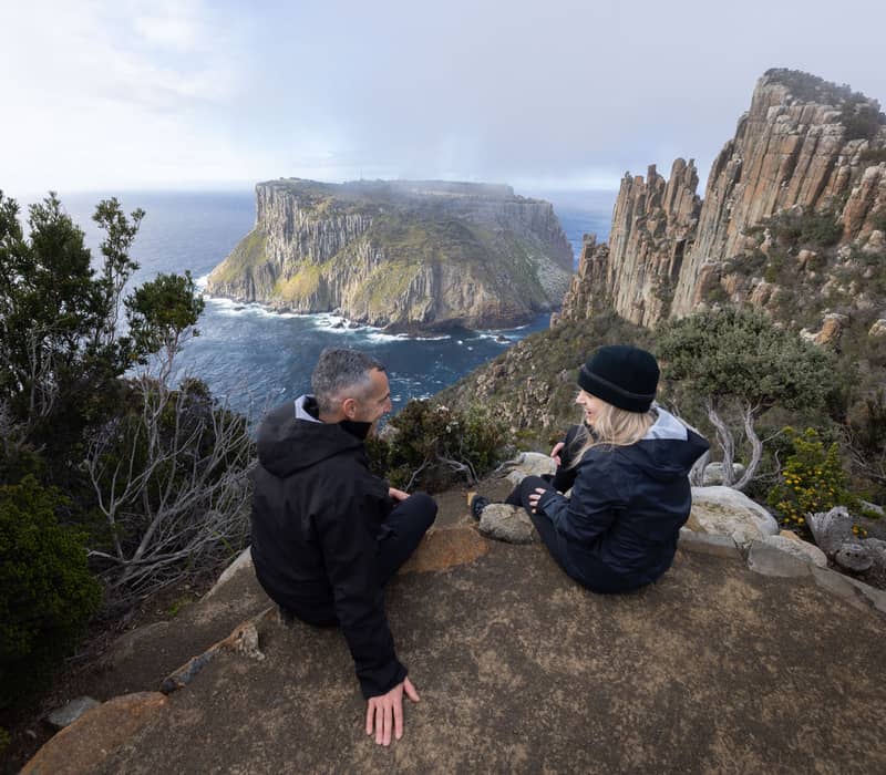 Two people sit on a rocky cliff edge with a view of a massive sea stack in the distance.