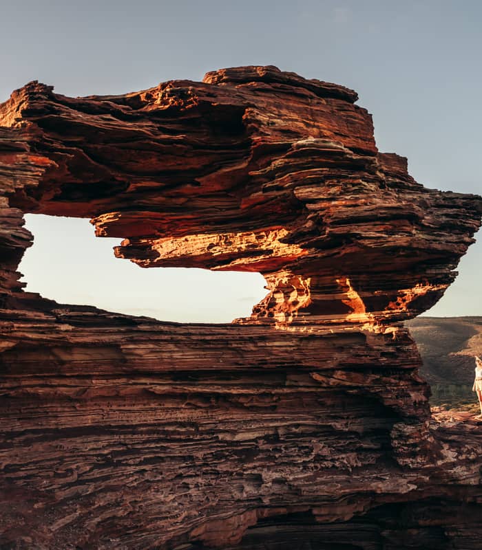 A hiker stands beside Nature's Window, a natural rock arch that perfectly frames the Murchison River winding through the gorge below.