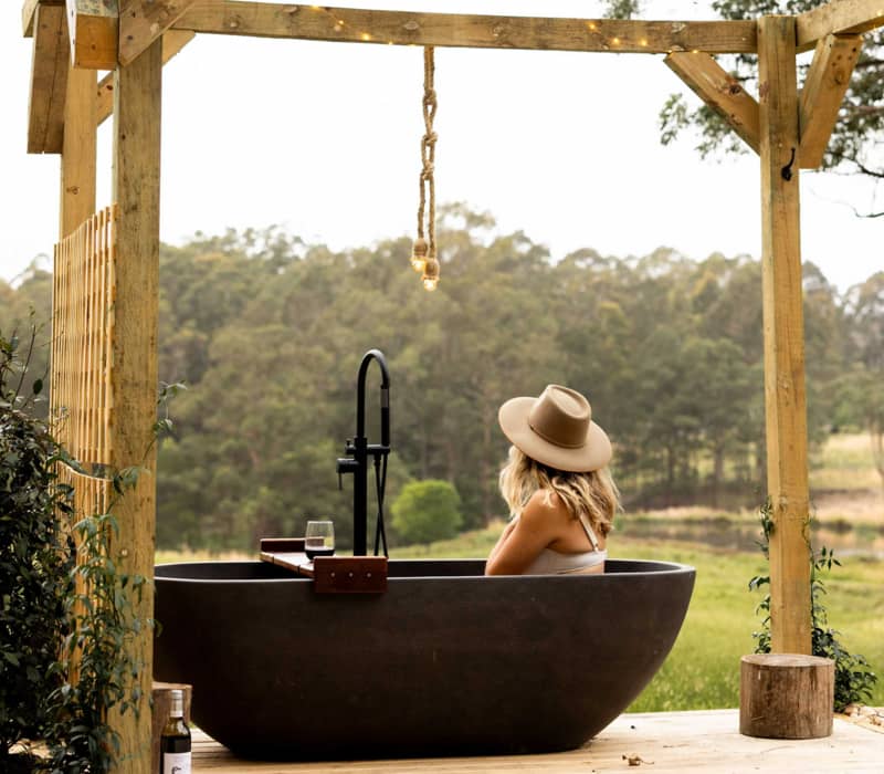 A woman in a tan wide-brimmed hat relaxes in a dark outdoor stone bathtub on a rustic wooden deck, looking out over a serene, lush green valley at a peaceful bush retreat.