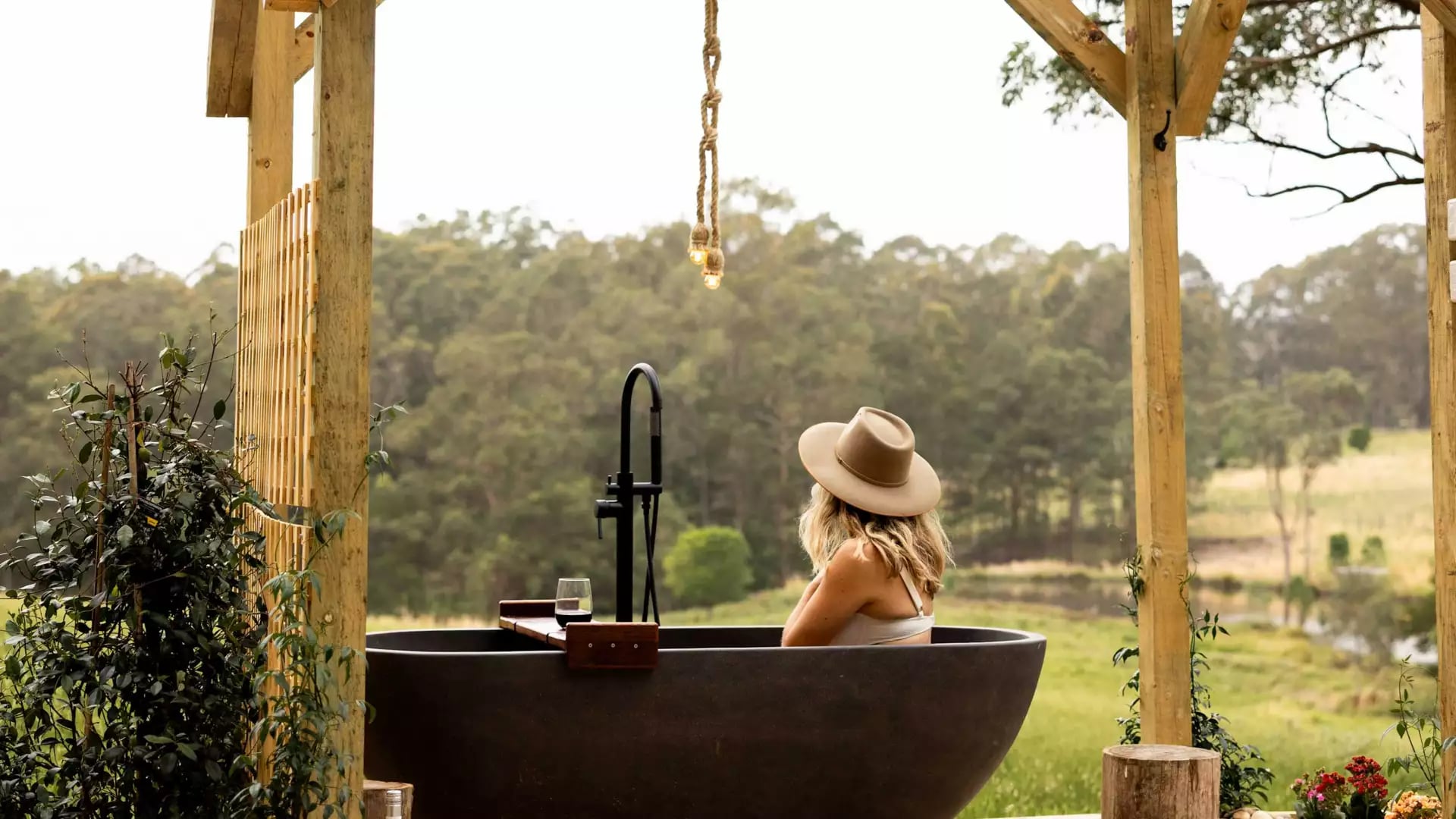 A woman in a tan wide-brimmed hat relaxes in a dark outdoor stone bathtub on a rustic wooden deck, looking out over a serene, lush green valley at a peaceful bush retreat.