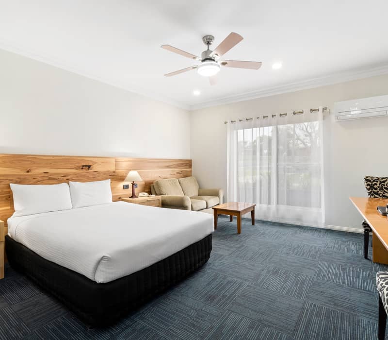 Modern two-storey hotel suite with a white queen bed, light grey leather sofa, and wooden stairs next to a small kitchenette.