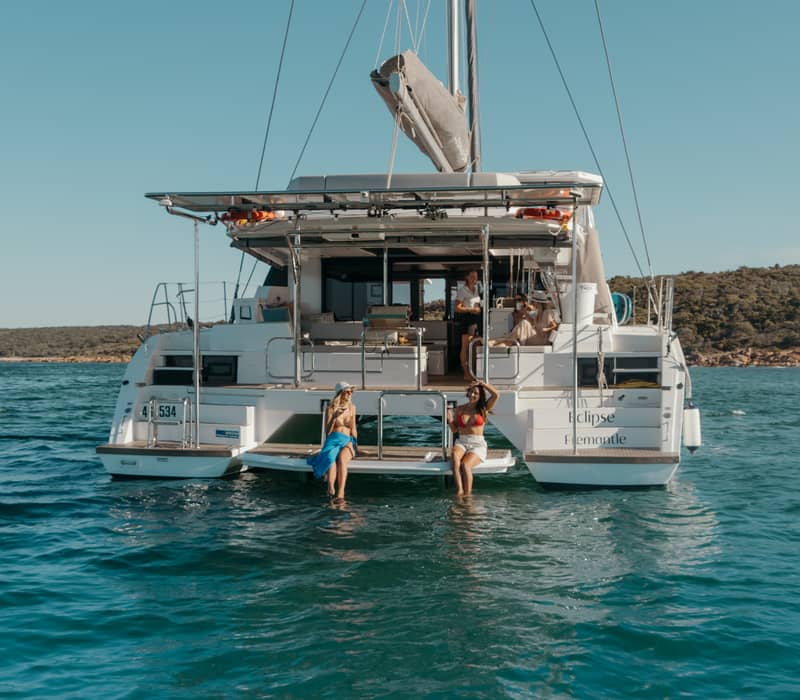 Two women sitting on the back of the Eclipse Expeditions catamaran with their feet in the water at Meelup Beach.