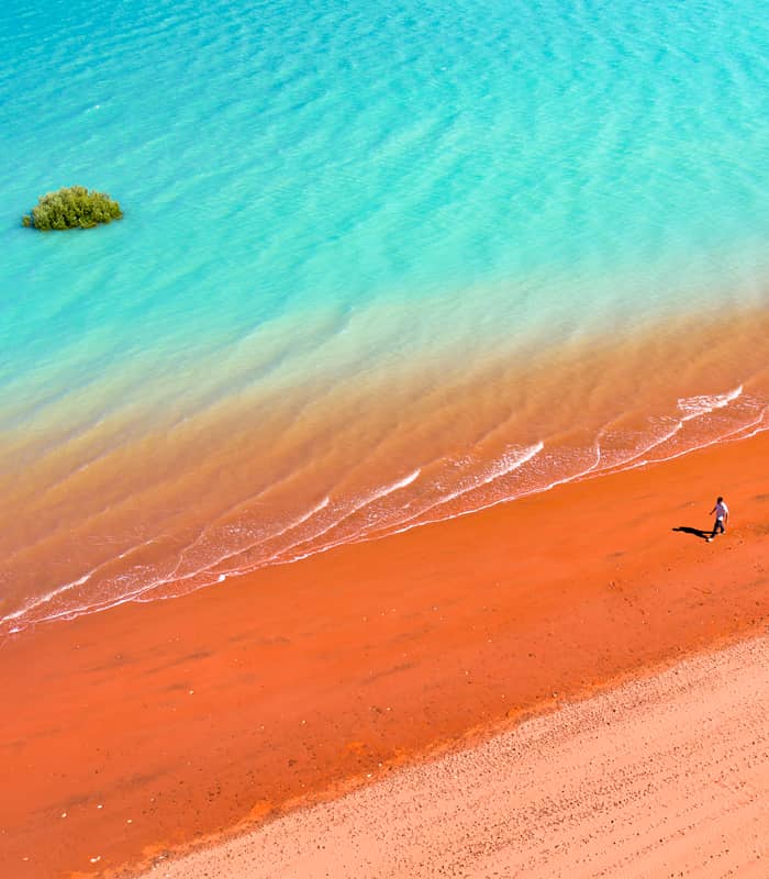 High-angle shot of a person walking on a bright red sandy beach beside clear turquoise ocean water.