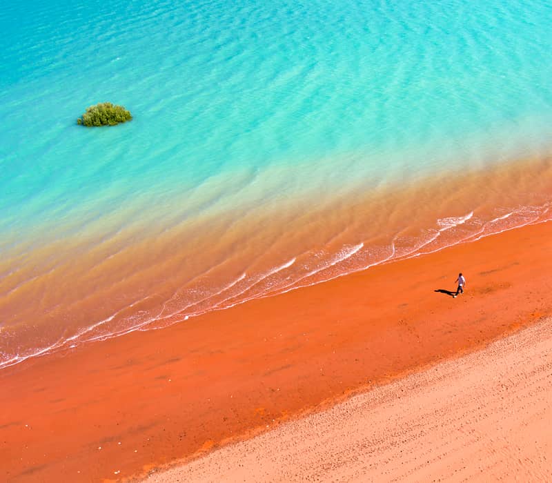 High-angle shot of a person walking on a bright red sandy beach beside clear turquoise ocean water.