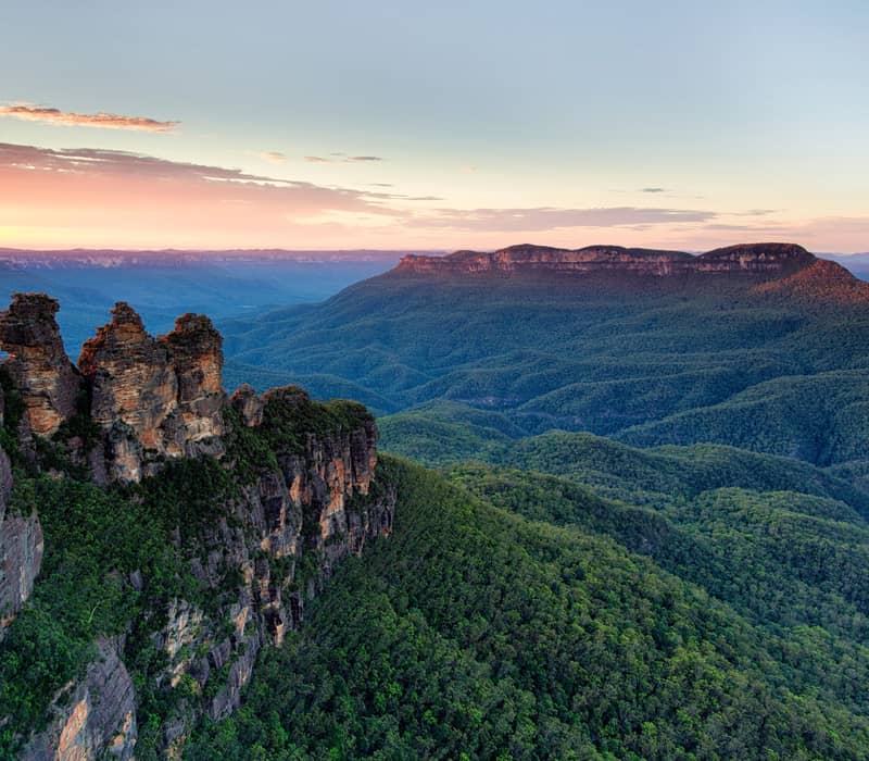 A panoramic sunset view of the Blue Mountains ranges with the Three Sisters rocks silhouetted against a pink sky.
