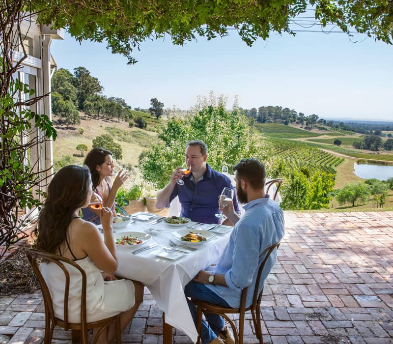 Four people dining outdoors at a white-clothed table with a scenic view of Hunter Valley vineyards.