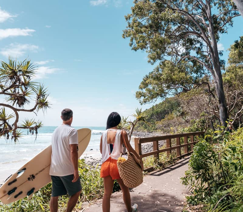 A man carrying a surfboard and a woman walking along a paved coastal path next to a rocky beach.