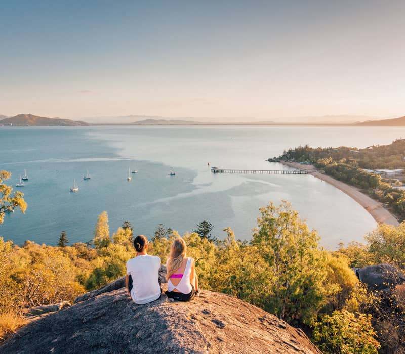 Couple sitting on a large rock overlooking a tropical bay with boats at sunset.