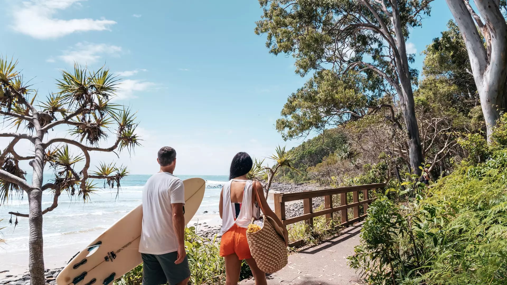 A man carrying a surfboard and a woman walking along a paved coastal path next to a rocky beach.