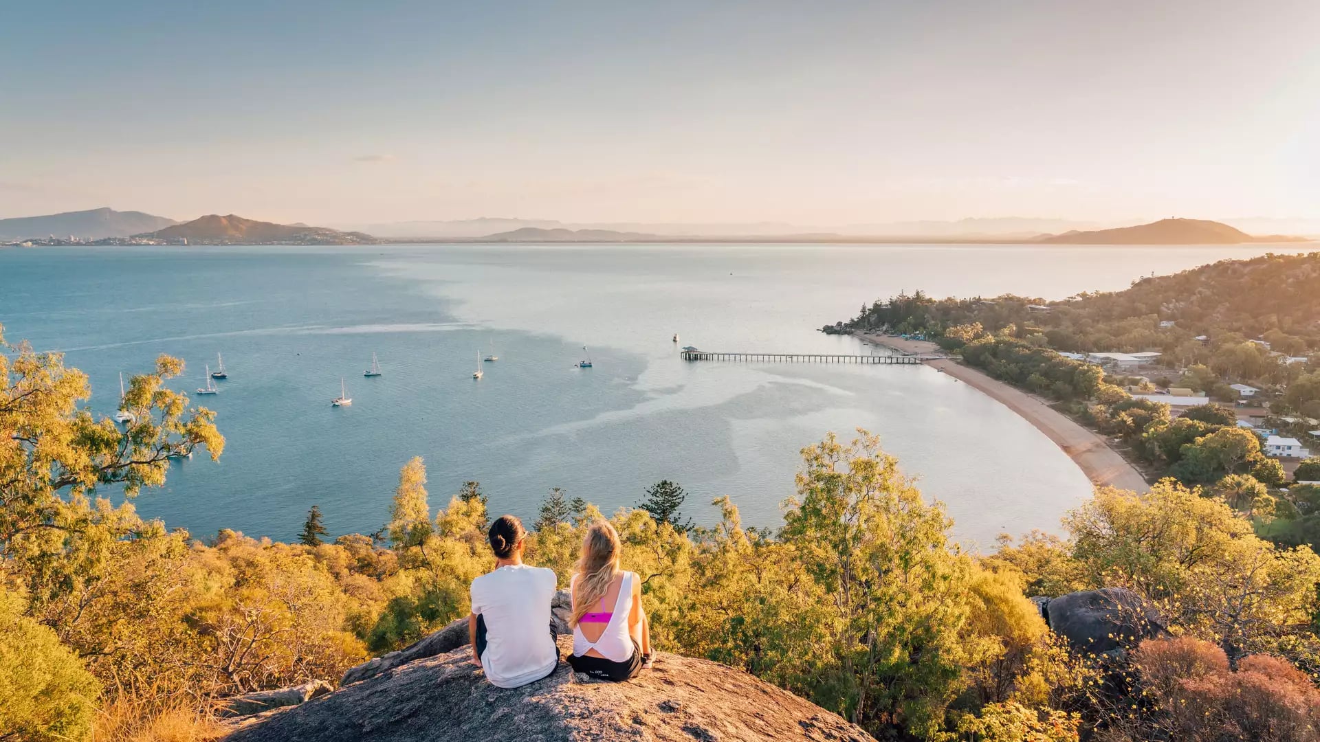 Couple sitting on a large rock overlooking a tropical bay with boats at sunset.
