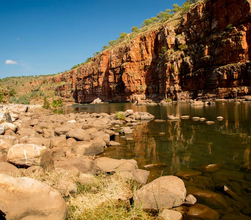 High red rock walls of a gorge reflecting in a river with boulders in the foreground, The Kimberley, WA.