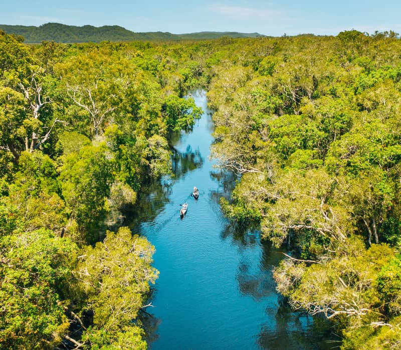 Drone photography of two canoes on a narrow blue river winding through thick Australian bushland.