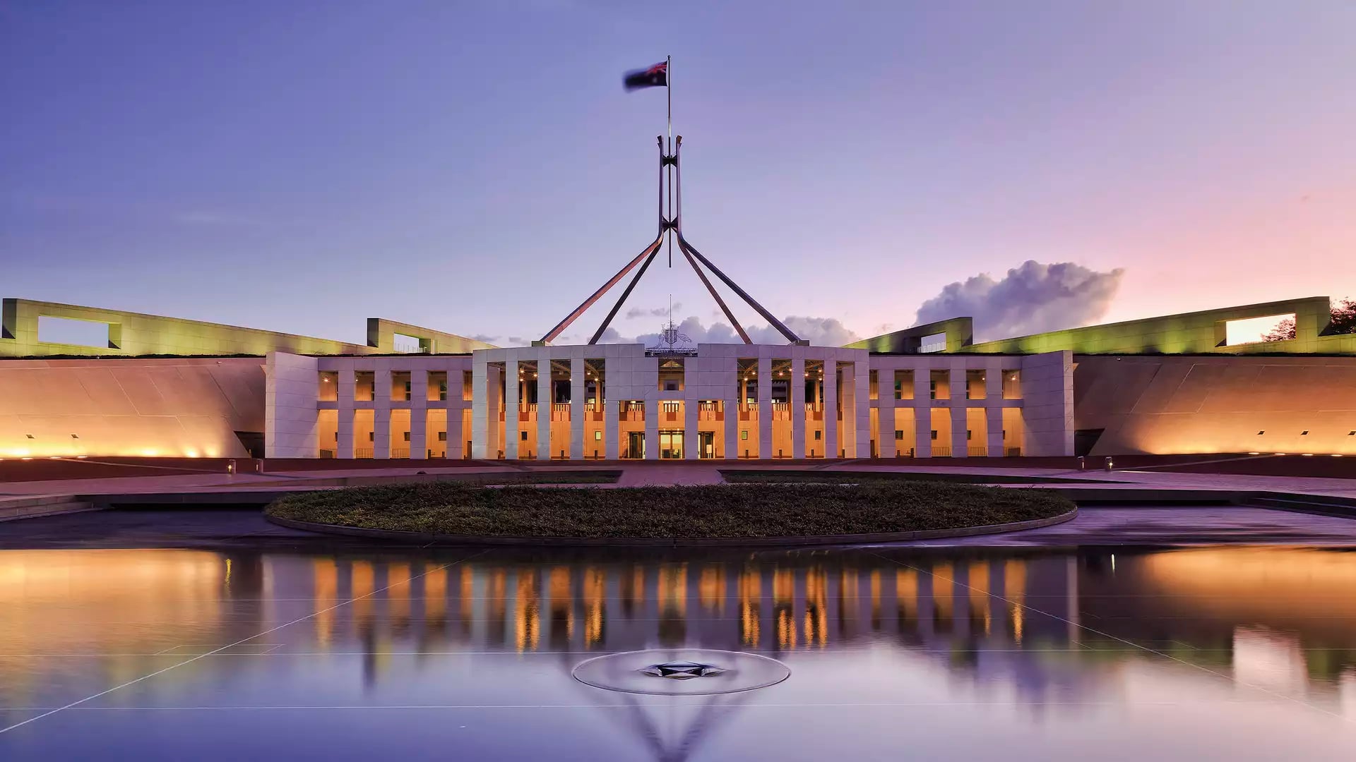 The facade of Parliament House in Canberra reflected in a still pool during a purple and orange sunset.