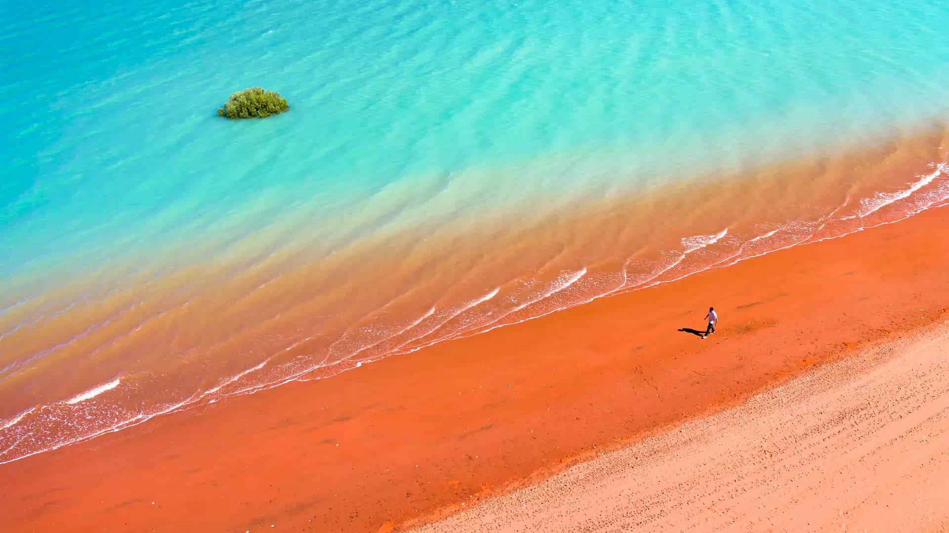 High-angle shot of a person walking on a bright red sandy beach beside clear turquoise ocean water.
