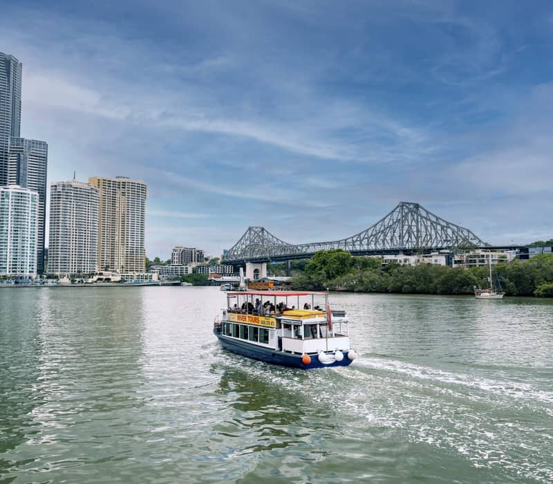 Sightseeing ferry on a river with the Story Bridge and city skyscrapers in the background.