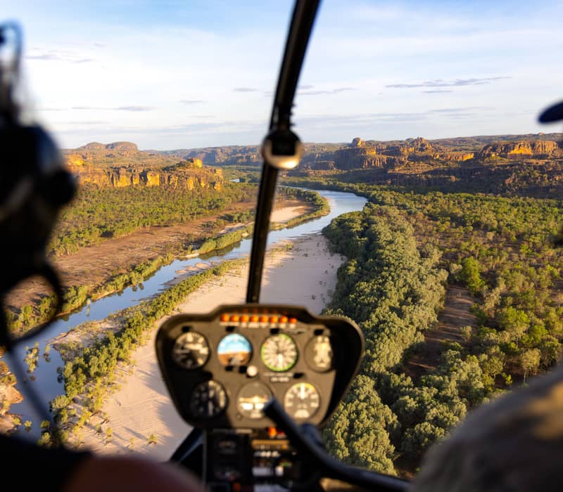 View from inside a helicopter cockpit looking out over a wide, sandy riverbed winding through a lush green forest toward distant orange sandstone cliffs at sunset.