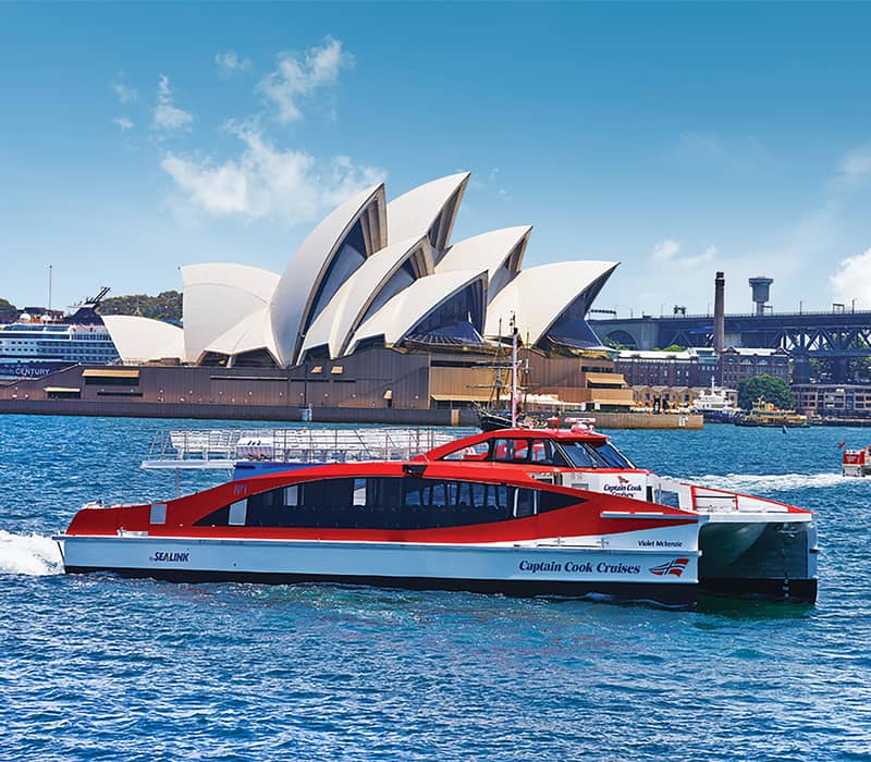 A Captain Cook Cruises ferry navigating blue waters in front of the Sydney Opera House under a bright sky.