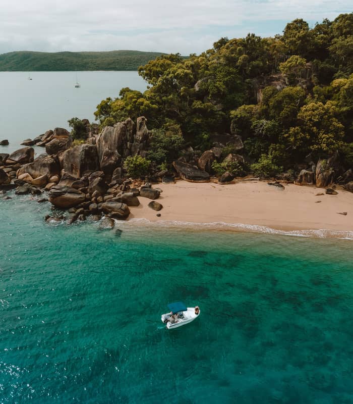 An aerial perspective shows a small white boat anchored in turquoise water next to a pristine sandy cove framed by rocky cliffs and lush green forest on Orpheus Island.