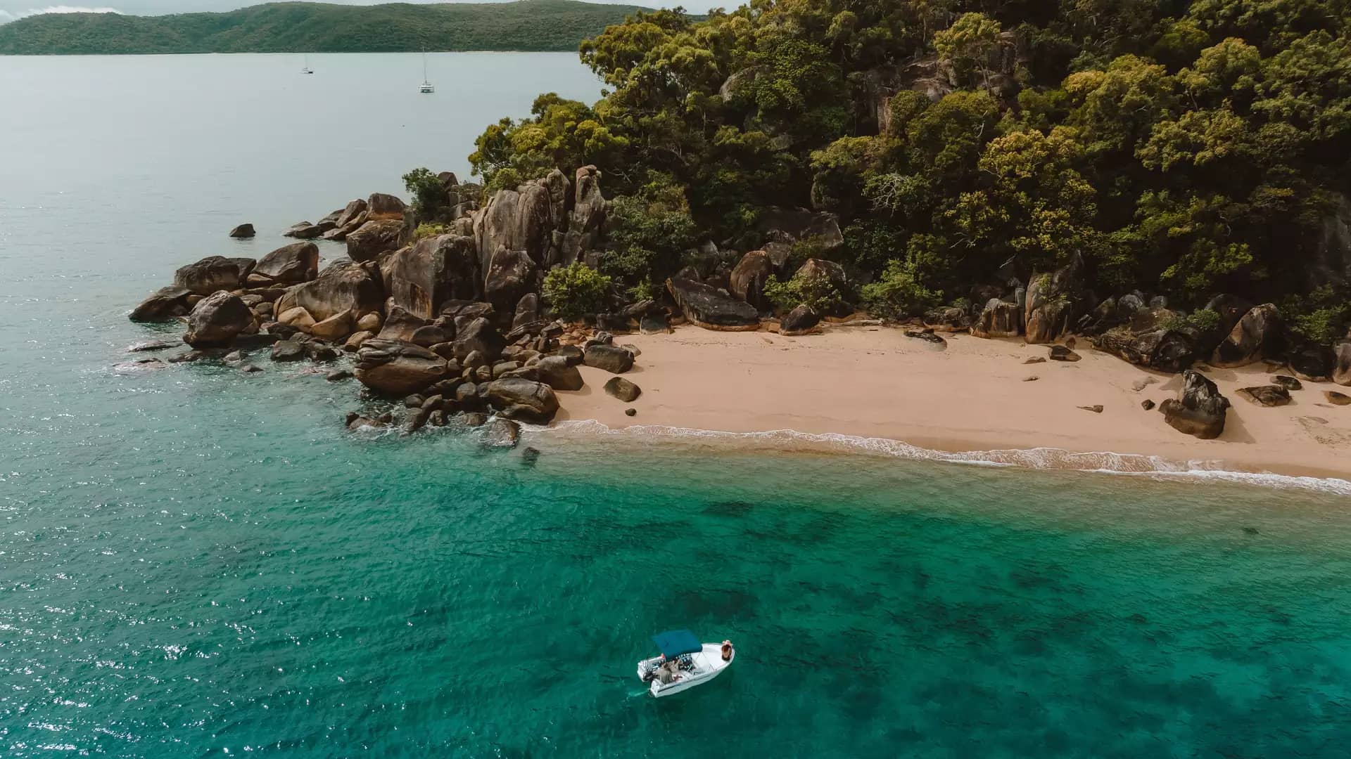 An aerial perspective shows a small white boat anchored in turquoise water next to a pristine sandy cove framed by rocky cliffs and lush green forest on Orpheus Island.