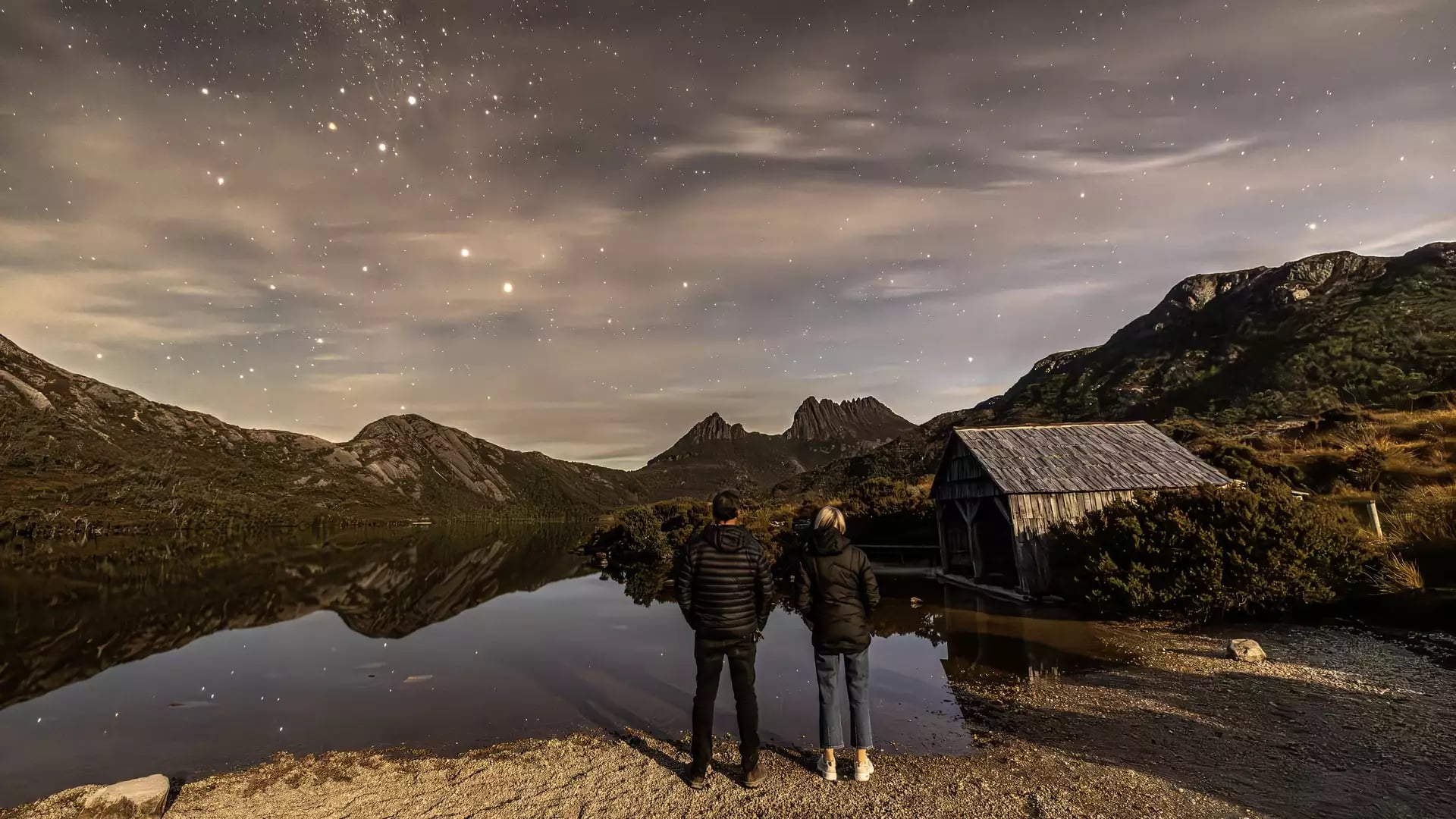 Rear view of a man and woman in winter jackets standing on a pebbled lake shore at night under a starry sky next to a small wooden hut.