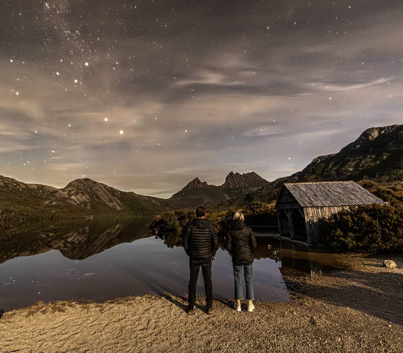 Rear view of a man and woman in winter jackets standing on a pebbled lake shore at night under a starry sky next to a small wooden hut.