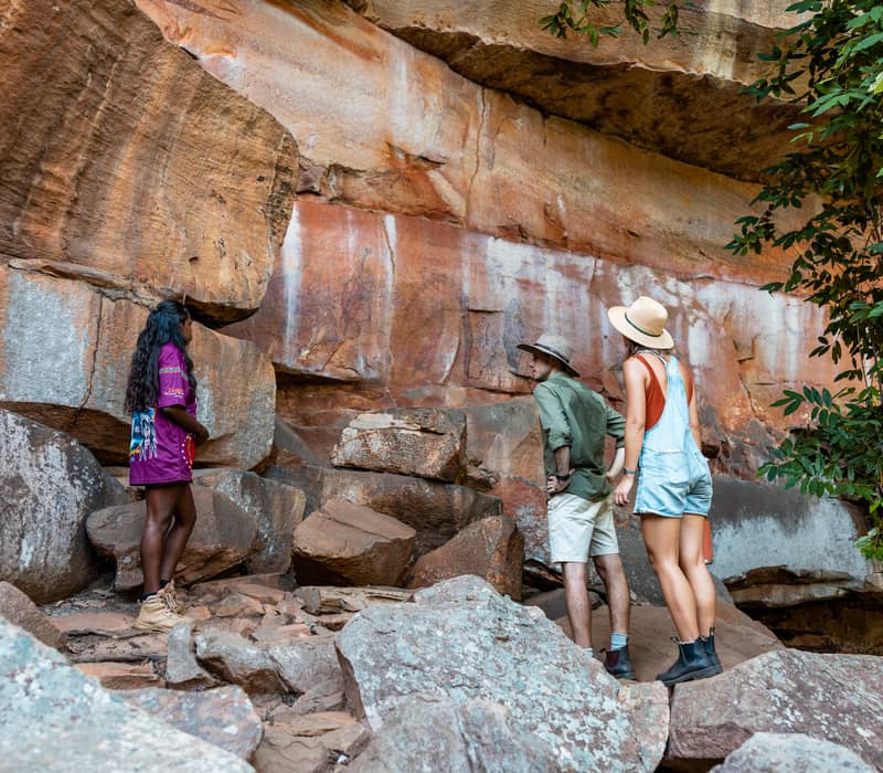 Two tourists and an Indigenous guide look up at a high, weathered rock wall with visible ancient paintings.