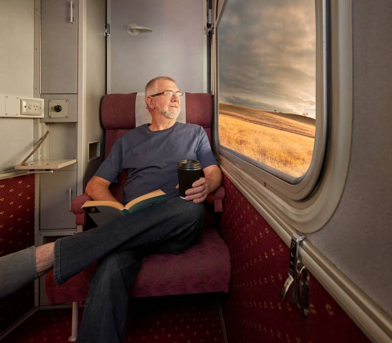 A man sits comfortably in a train sleeper compartment with a book and coffee, gazing out at the golden outback landscape.