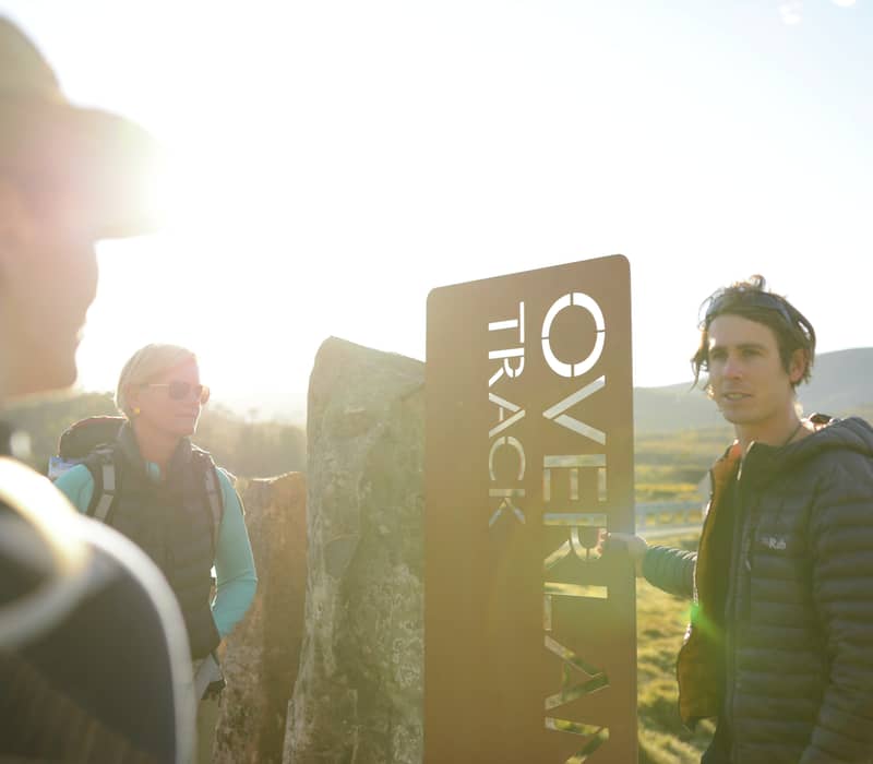 A guide talking to hikers next to the Overland Track sign at sunset in Tasmania.