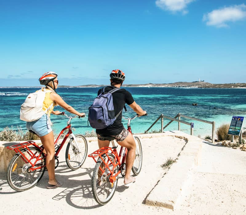 Two people on red bicycles looking out over a stunning turquoise bay from a paved path.