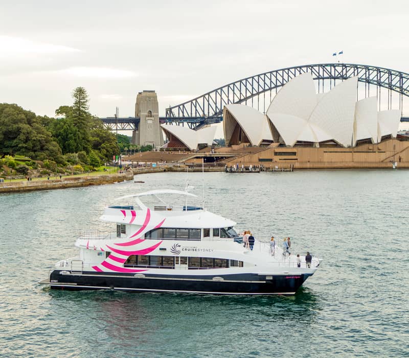 A white and pink branded cruise boat sailing on Sydney Harbour with the Opera House in the background.