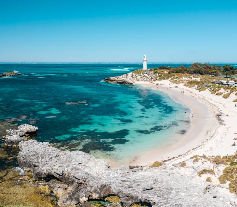 Wide aerial view of a white sandy beach, turquoise water, and a white lighthouse on a grassy headland.