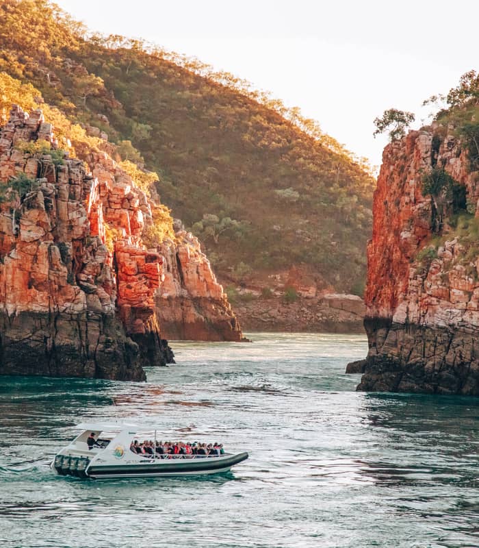 A white tour boat carrying passengers through a narrow turquoise water gap between towering red rock cliffs.