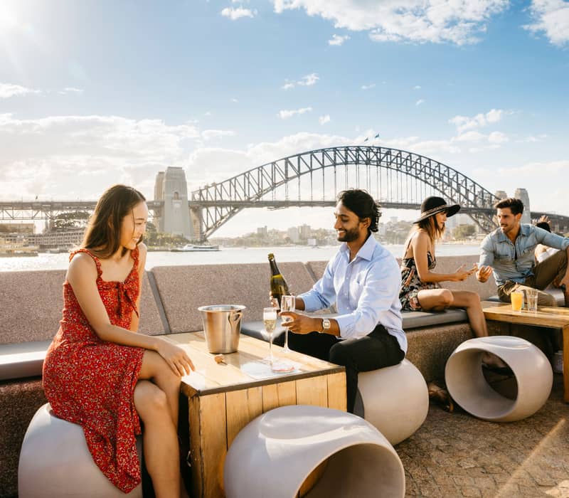 A diverse group of friends sitting at an outdoor bar with the Sydney Harbour Bridge in the background.