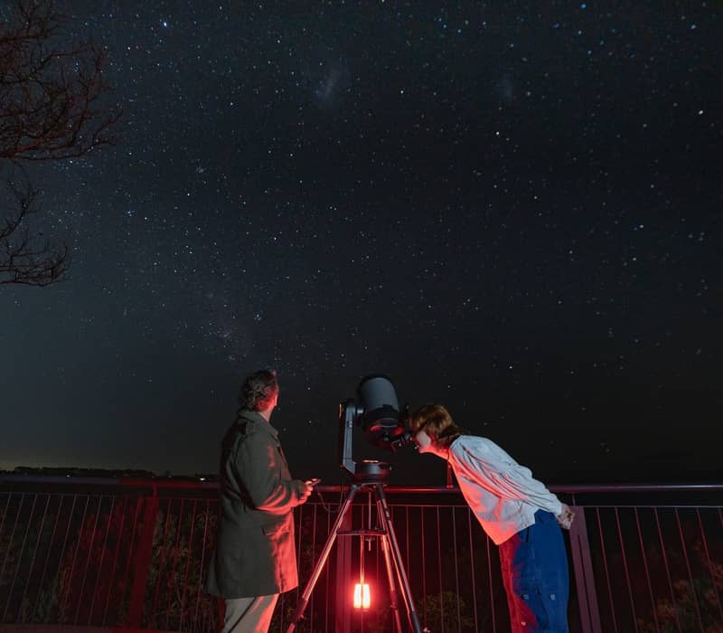 Two people using a large telescope to view a starry night sky on an outdoor platform.