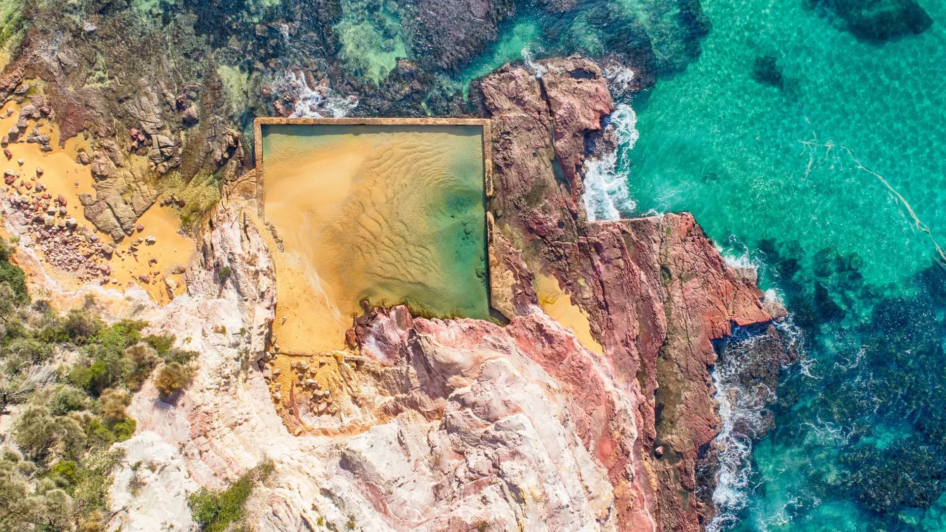 Aerial perspective of a rectangular man-made ocean pool surrounded by rugged red rocks.