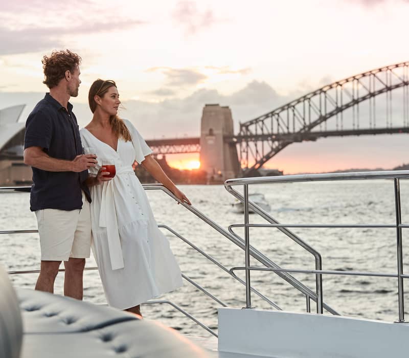 Couple standing on a boat deck holding drinks with the Sydney Harbour Bridge silhouetted against a sunset sky.