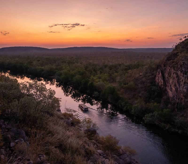 A serene evening view from an elevated vantage point showing a tour boat navigating the Katherine River as the sky transitions into deep twilight orange.