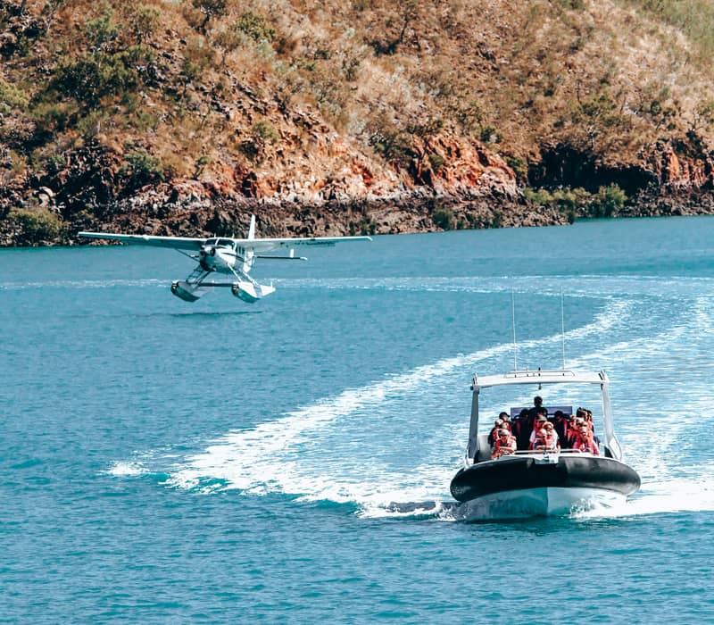 A seaplane landing on the water while a tour boat filled with passengers passes nearby in the Kimberley.