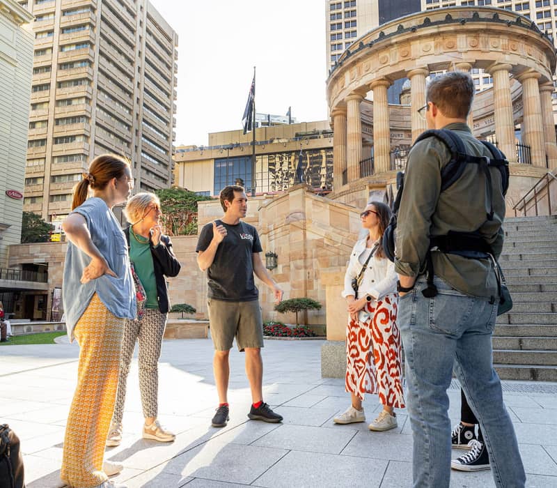 Travelers standing in a public square listening to a guide in front of a circular stone monument with pillars in Brisbane.