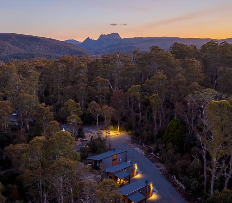 Aerial twilight view of lit-up modern cabins surrounded by thick green forest with a mountain range in the distance under a sunset sky.