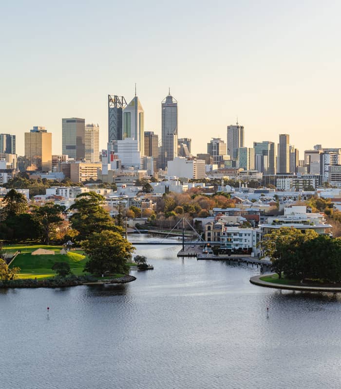 A high-angle aerial shot of the Perth city skyline with tall office buildings under a pale evening sky. In the foreground, the dark blue Swan River winds past green parks and residential areas with several small bridges.