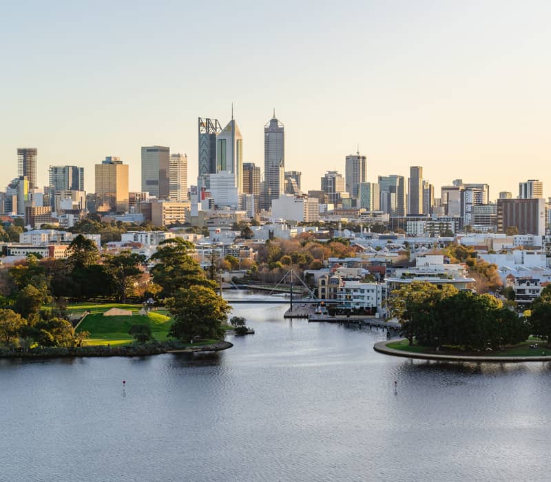 A high-angle aerial shot of the Perth city skyline with tall office buildings under a pale evening sky. In the foreground, the dark blue Swan River winds past green parks and residential areas with several small bridges.