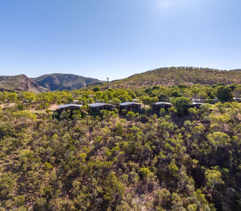 An aerial panorama of the Discovery Park with white-roofed buildings nestled among green trees and arid, hilly scrubland next to a wide, blue river that is flanked by a massive, sheer red rock cliff.