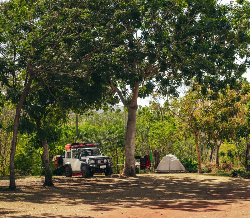 A white 4WD vehicle and a small grey tent are parked in the shade of large, leafy trees on a dirt campsite at a caravan park.