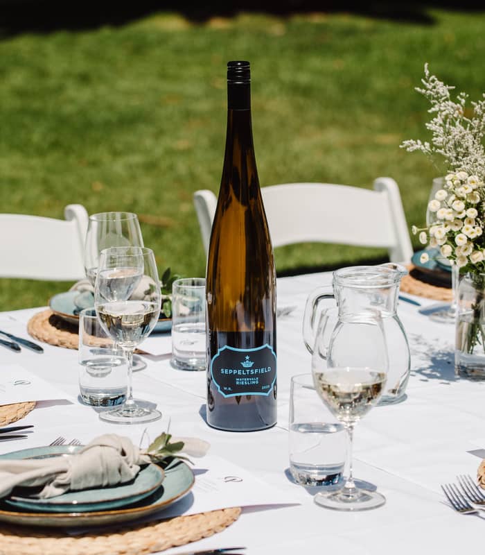 A bright, outdoor dining table set with white linen, featuring a bottle of Seppeltsfield Riesling, wine glasses, and a native Australian flower arrangement with a Protea.