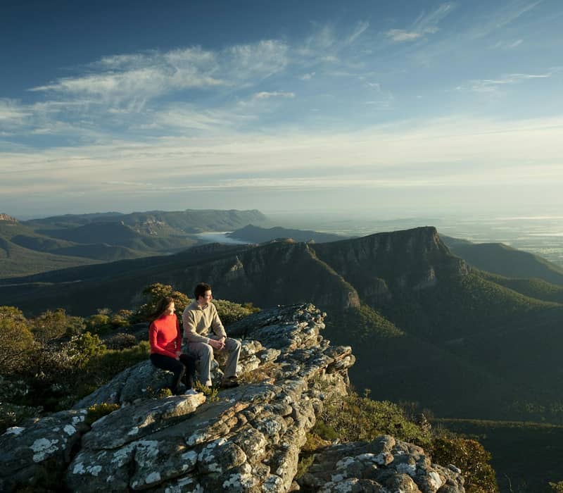 Couple sitting on rocky ledge overlooking expansive mountain landscape in the Grampians.