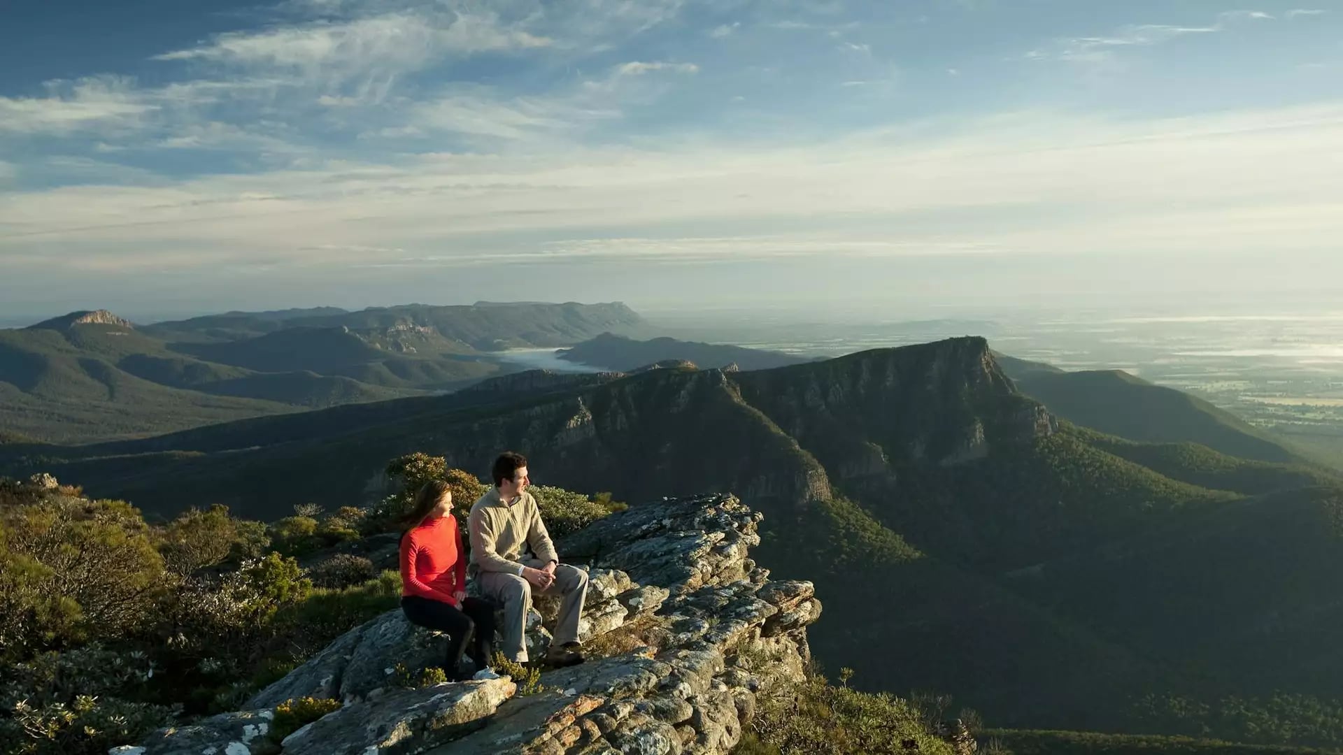 Couple sitting on rocky ledge overlooking expansive mountain landscape in the Grampians.