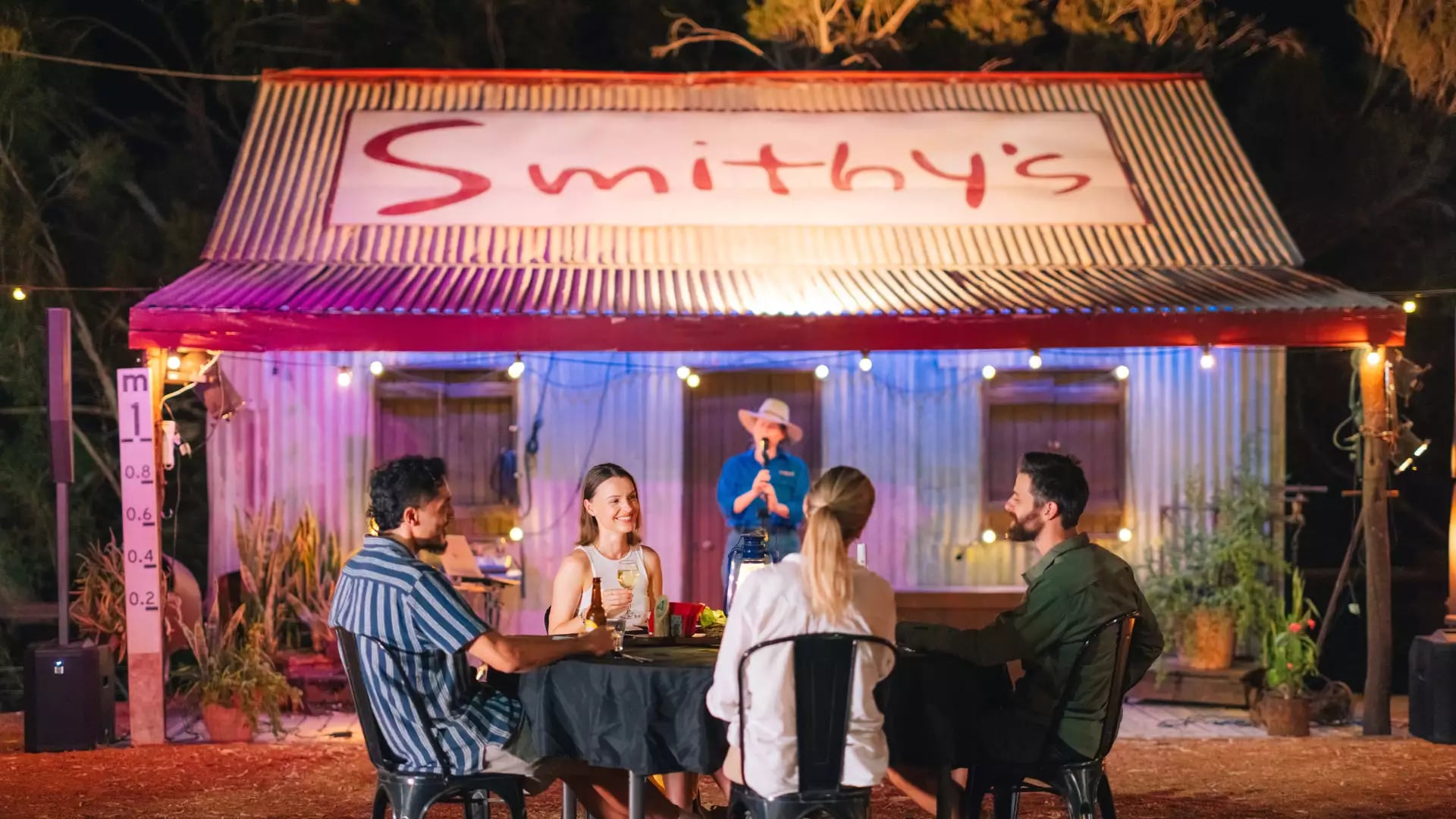 Four people sitting at a dinner table outside a rustic corrugated iron building with "Smithy's" sign at night.